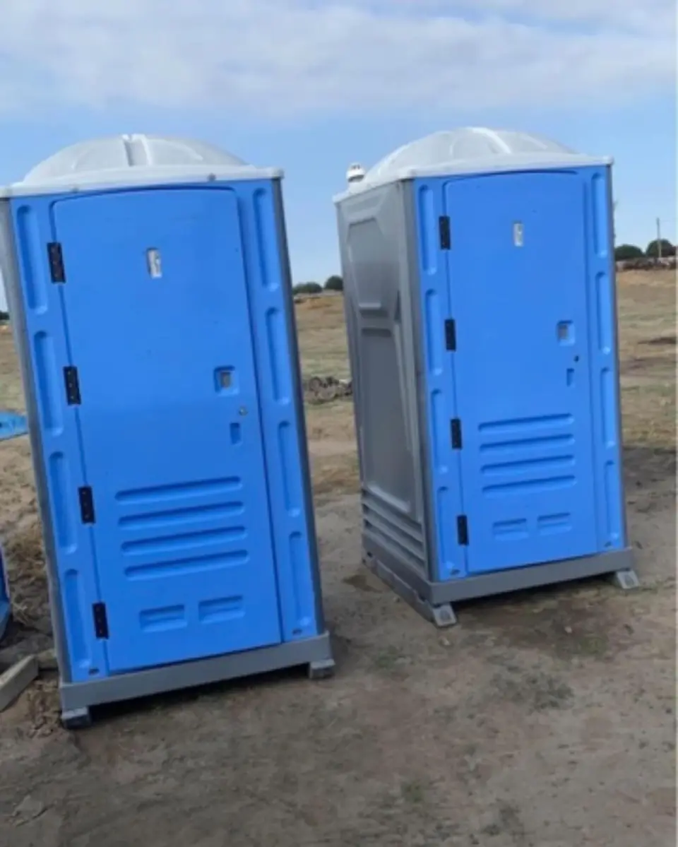 Two blue standard porta potties side by side on open ground